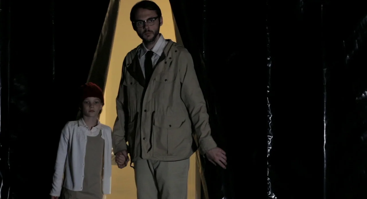 Sound Of My Voice (2011) movie still: fullbody — A man and a child emerge from a dark space into a bright area. They stand in front of black plastic…