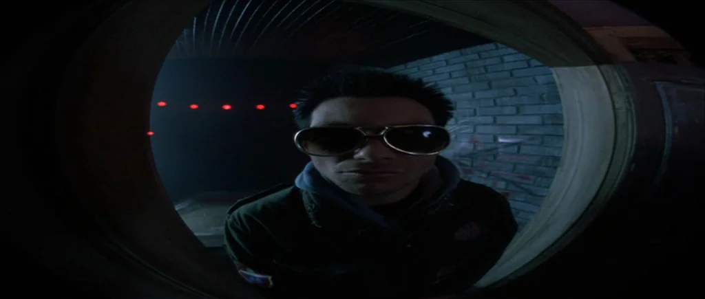 True Romance (1993) movie still: closeup — A dark brick exterior wall is framed through a circular fisheye door peephole. A man stares directly into…