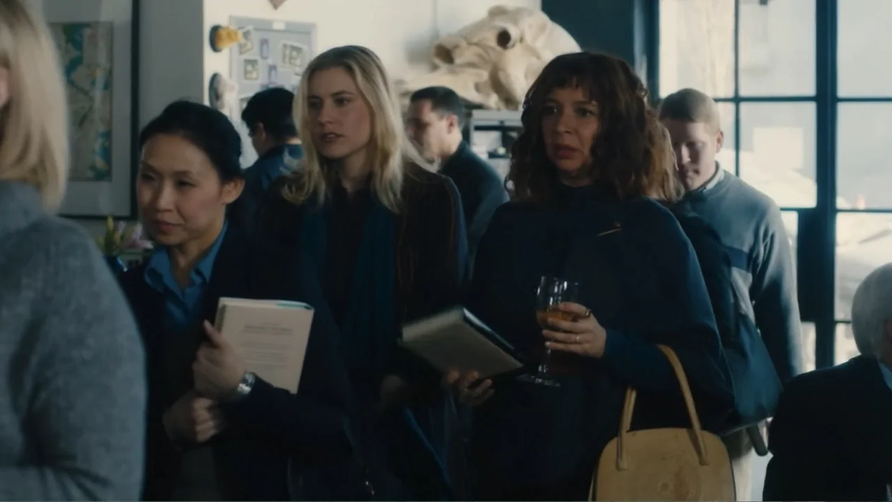 Maggie's Plan (2016) movie still: medium — A crowded interior space with artwork on the walls and a large window. Three women stand in a cluster…