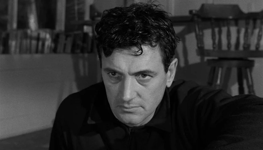 Seconds (1966) movie still: medium — A man stands in a dimly lit room with wooden furnishings and books visible. The man stares directly forward…