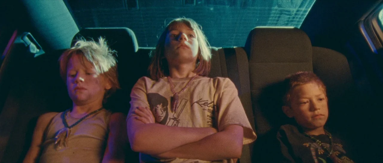 Riddle of Fire (2023) movie still: medium — The interior of a car at night illuminated by external blue and warm light. Three children sit across…