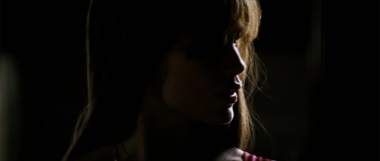 Elektra (2005) movie still: closeup — A dark indoor space with a single light source striking one side of a face. A woman with long hair faces away…