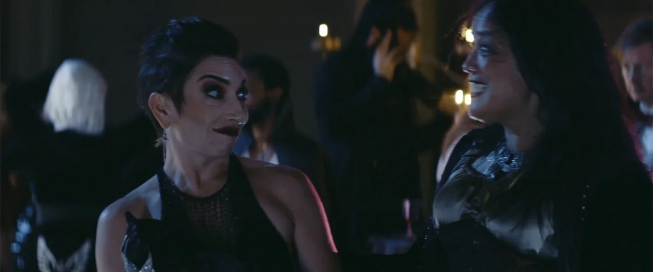 Mennonite's Bite (2023) movie still: medium — A dimly lit ballroom features blurred figures in the background under soft warm light. Two women…