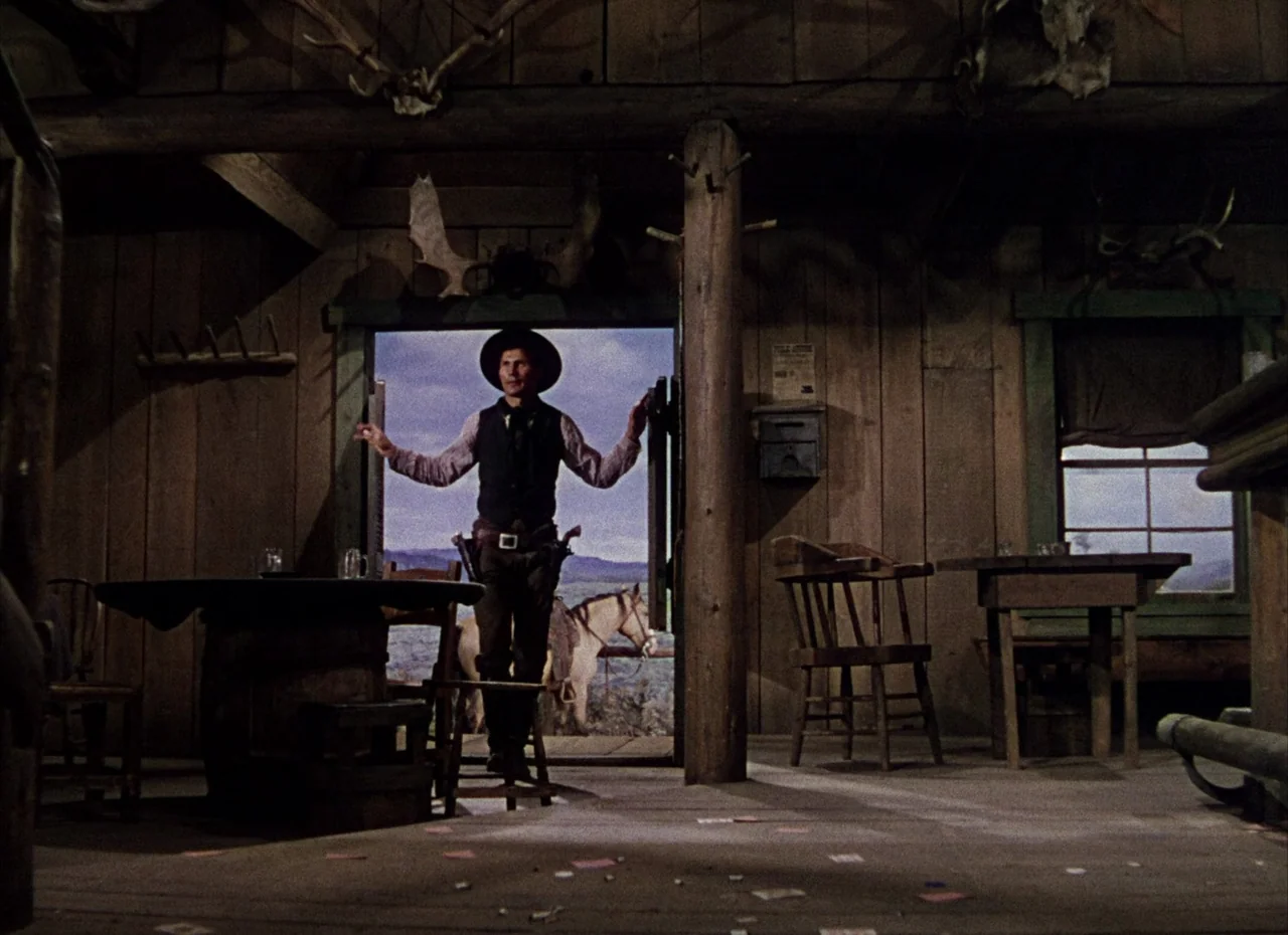 Shane (1953) movie still: fullbody — A rustic wooden building with antler decorations above the doorway and scattered playing cards. A man stands in…