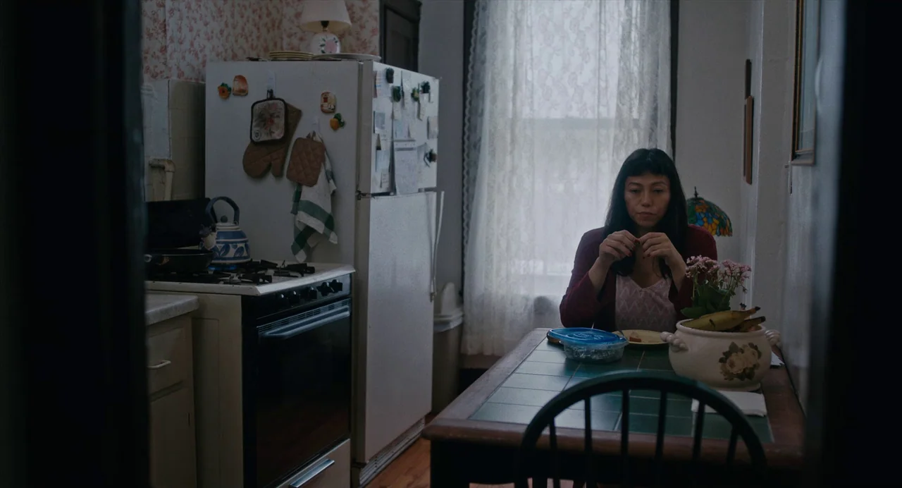 Lingua Franca (2019) movie still: medium — A residential kitchen features white appliances, patterned wallpaper, and a tile-top wooden dining table.…