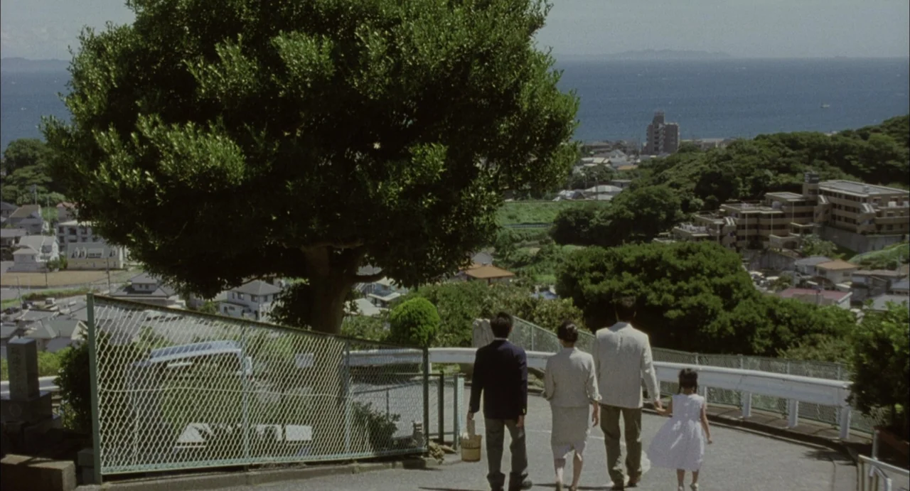 Still Walking (2008) movie still: wide — A group of four people walks down a road overlooking a coastal town. The path features a chain-link fence…
