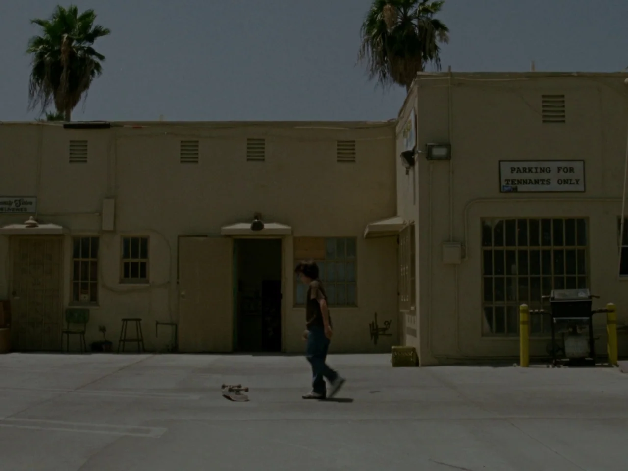 Mid90s (2018) movie still: fullbody — A beige-colored apartment complex building stands behind a concrete parking area under daylight. A teenage boy…