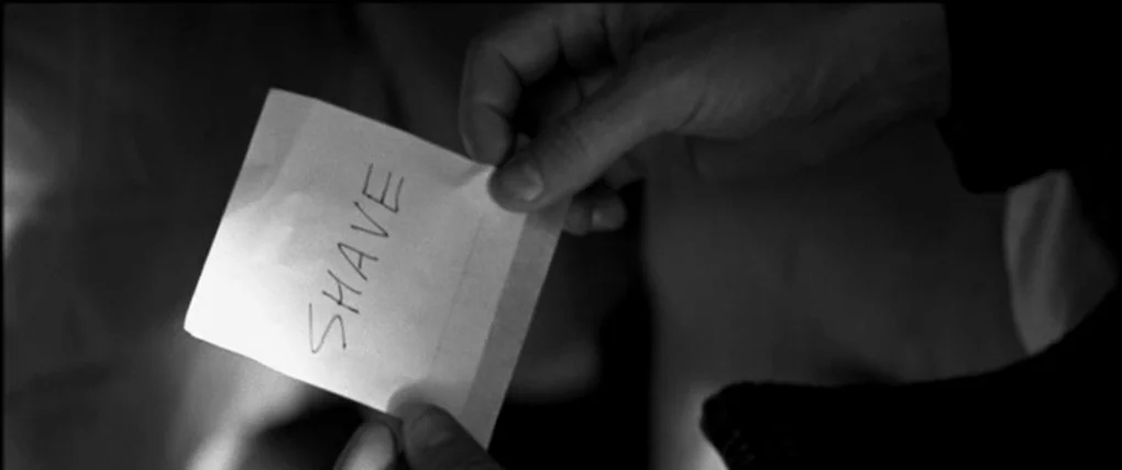 Memento (2000) movie still: closeup — A dark, dimly lit environment provides the backdrop for a small white note. Handheld paper with the word SHAVE…
