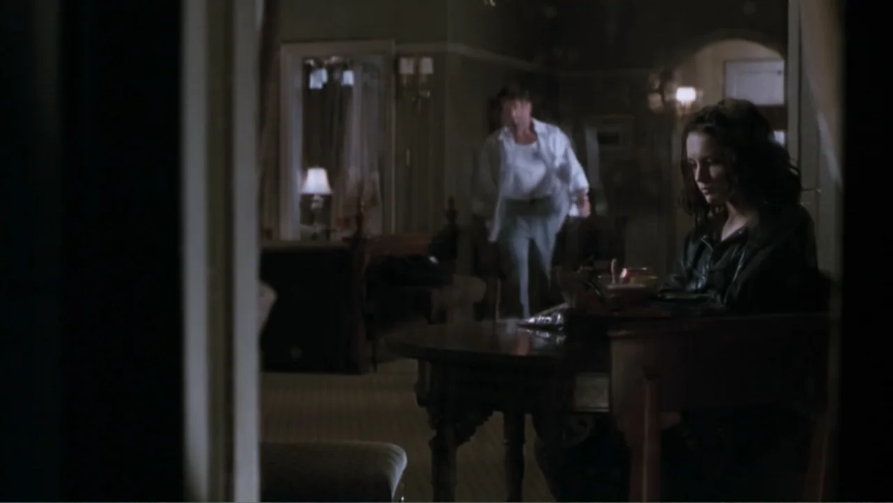 Get Carter (2000) movie still: medium — A dimly lit room features a wooden table where a woman sits in the foreground. A man walks through the…