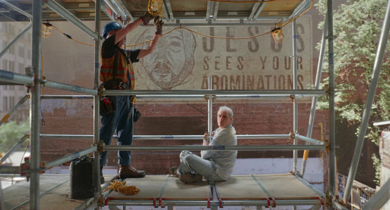 Beau Is Afraid (2023) movie still: fullbody — Two men stand and sit on metal scaffolding mounted against a large urban brick wall. A mural of a face…