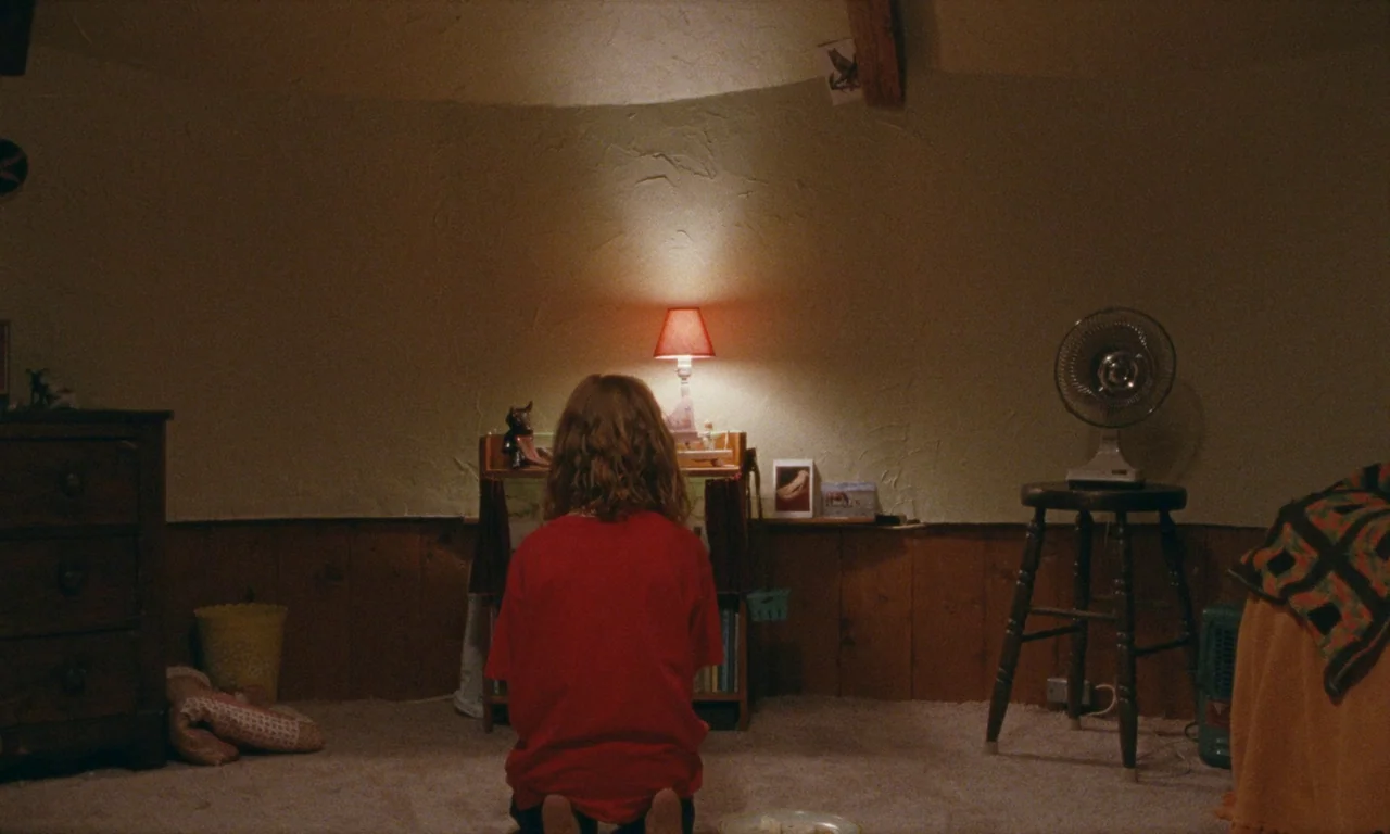 Janet Planet (2023) movie still: medium — A dimly lit room features wood-paneled walls and a beige carpeted floor. A child in a red shirt kneels on…