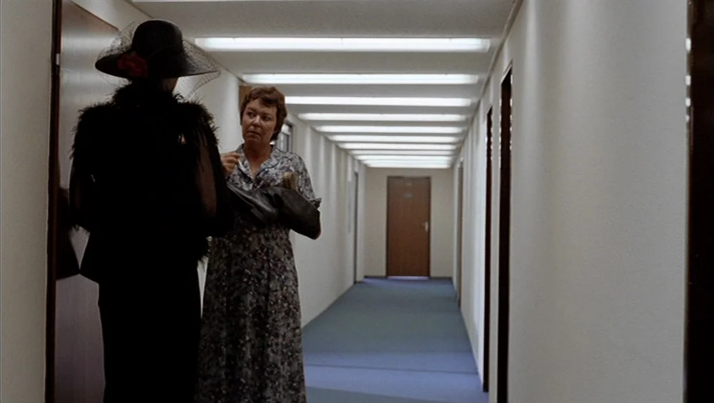 In A Year Of Thirteen Moons (1978) movie still: medium — A long, sterile building corridor with blue carpeting and white walls features recessed…