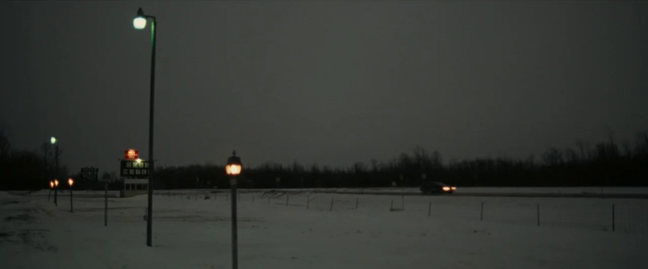 For Ellen (2012) movie still: establishing — A dark, snow-covered roadside landscape at night features rows of glowing streetlamps. A faint…