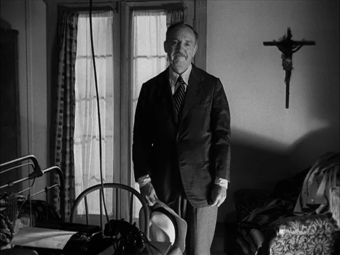 Ace In The Hole (1951) movie still: medium — A room contains a bed, a desk with a telephone, and a wall crucifix. An elderly man stands centrally in…