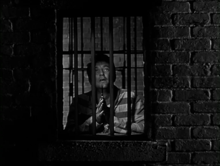 The Night Of The Hunter (1955) movie still: medium — A man stands behind iron vertical bars set into a textured brick wall surface. He holds his…