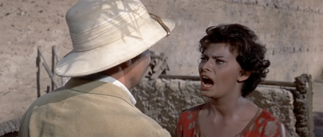 Legend of the Lost (1957) movie still: medium — A rural courtyard features rough-textured mud walls and simple wooden construction elements. A woman…