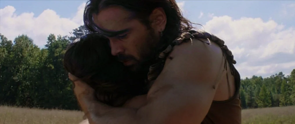 The New World (2005) movie still: medium — A man with long hair hugs a woman in an expansive, grassy field. The background features a thick line of…