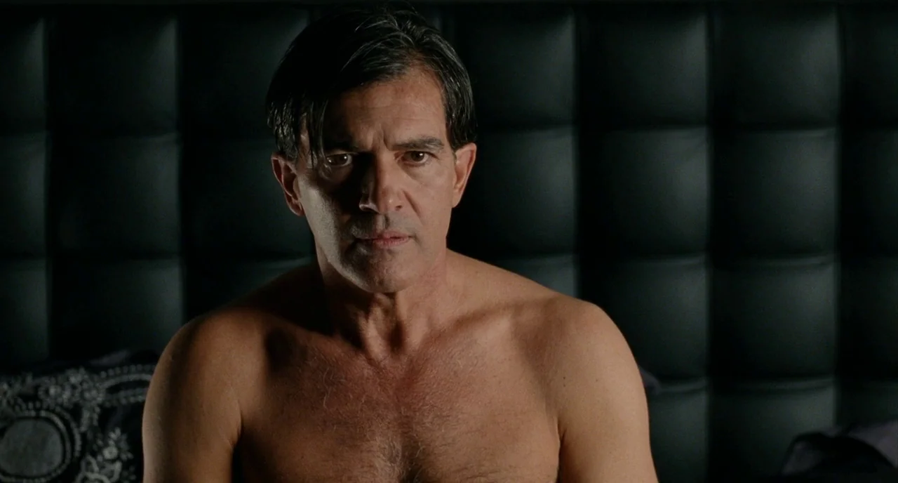 The Skin I Live In (2011) movie still: closeup — A man sits shirtless in front of a dark, tufted leather headboard. The subject stares forward with…