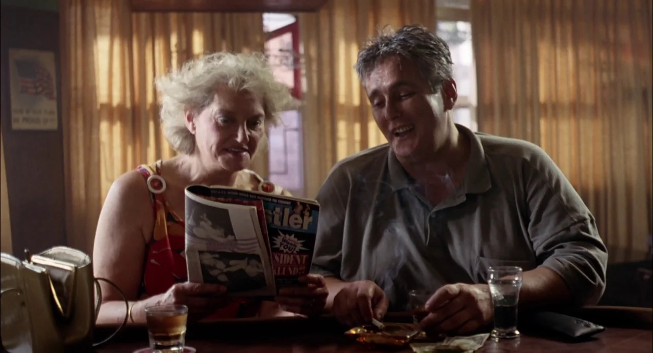Trees Lounge (1996) movie still: medium — A dimly lit diner booth interior with golden curtains covering a background window. An elderly woman reads…