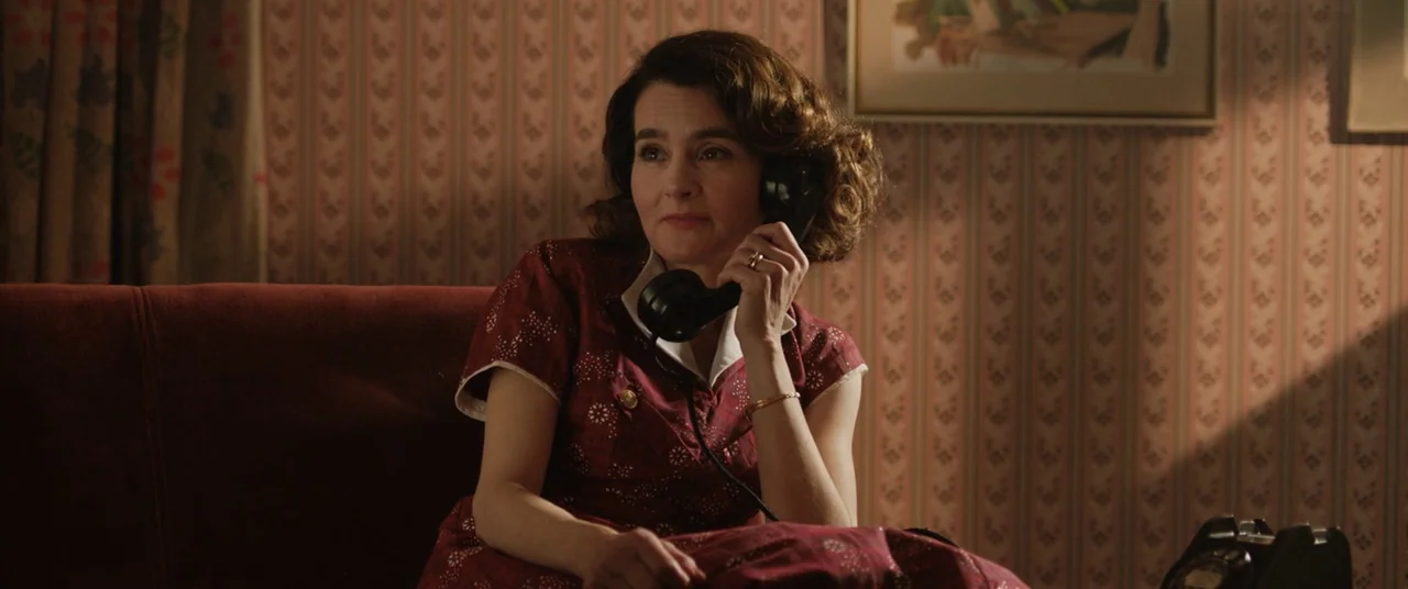 Domino (2005) movie still: medium — A 1950s living room features vertical striped wallpaper and a red velvet couch. A woman in a patterned maroon…