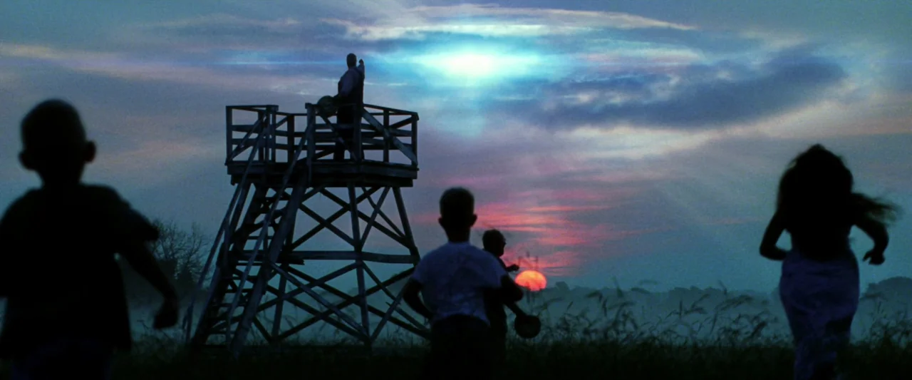 Armageddon (1998) movie still: wide — A field at twilight features a tall wooden lookout tower beneath a glowing sky. Several children run across…