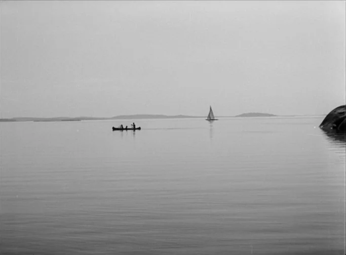 Summer Interlude (1951) movie still: wide — A vast, calm body of water spans the frame under an overcast sky. A small canoe and a sailboat travel…