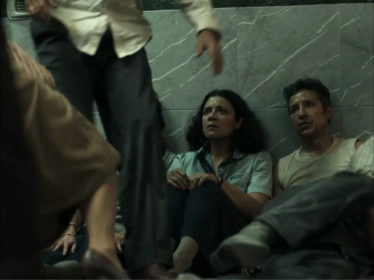 November (2025) movie still: medium — A cramped indoor hallway lined with grey marble-patterned tiles on the lower walls. People sit huddled on the…