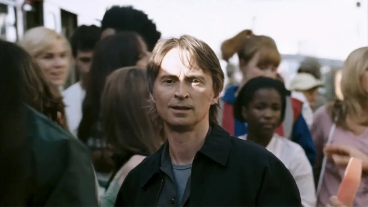 28 Weeks Later (2007) movie still: medium — A man stands in the center of a dense outdoor crowd during the day. He looks forward while surrounded by…