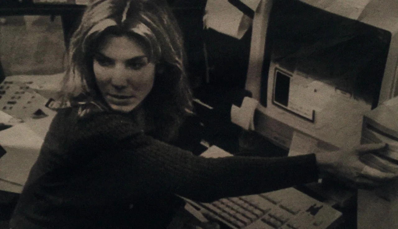Our Brand is Crisis (2015) movie still: medium — A dimly lit room contains a clunky desktop computer setup from the nineties. A woman looks over her…