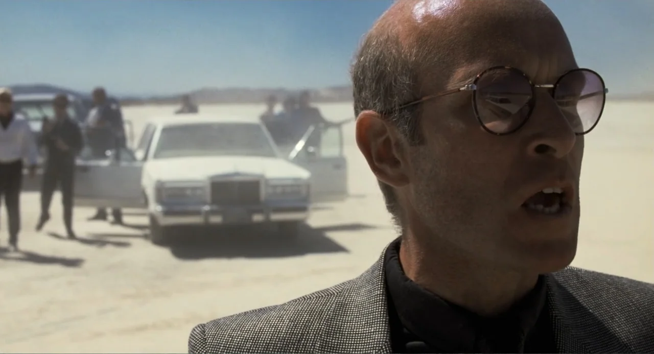 Lethal Weapon (1987) movie still: closeup — A vast, bright salt flat under a clear sky features a white luxury car. A man stands in the foreground…