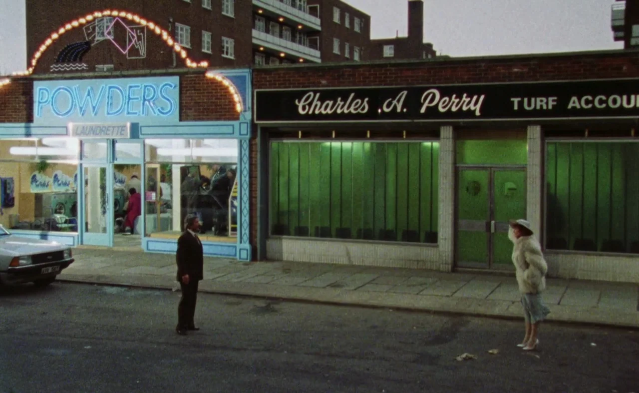 My Beautiful Laundrette (1985) movie still: wide — A row of two businesses with illuminated signs stands against a brick building at night. A man in…