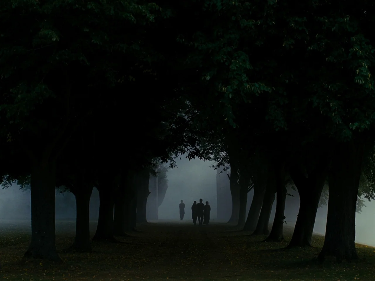 Saltburn (2023) movie still: wide — A dark, tree-lined path is filled with heavy ground fog during a dim day. Four individuals walk away from the…