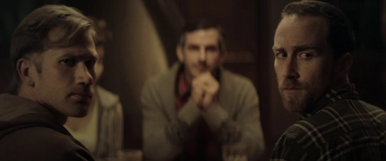 The Endless (2017) movie still: medium — A dimly lit indoor space with three men sitting at a table together. The men turn their heads to face…
