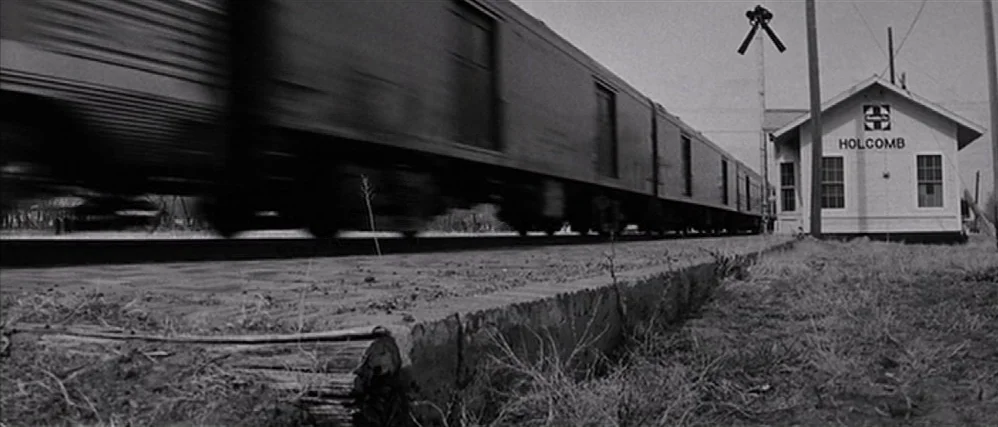 In Cold Blood (1967) movie still: wide — A small white train depot marked Holcomb stands near railroad tracks. A motion-blurred freight train…