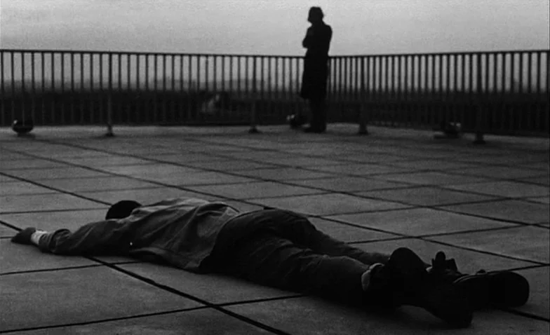 La Jetée (1962) movie still: wide — A flat concrete rooftop area features a simple railing along the perimeter edge. One figure lies prone on the…
