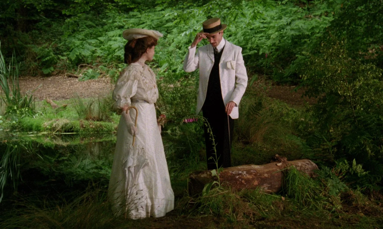 A Room With A View (1985) movie still: fullbody — A man and woman in formal Edwardian attire stand on the bank of a stream. The man tips his hat to…