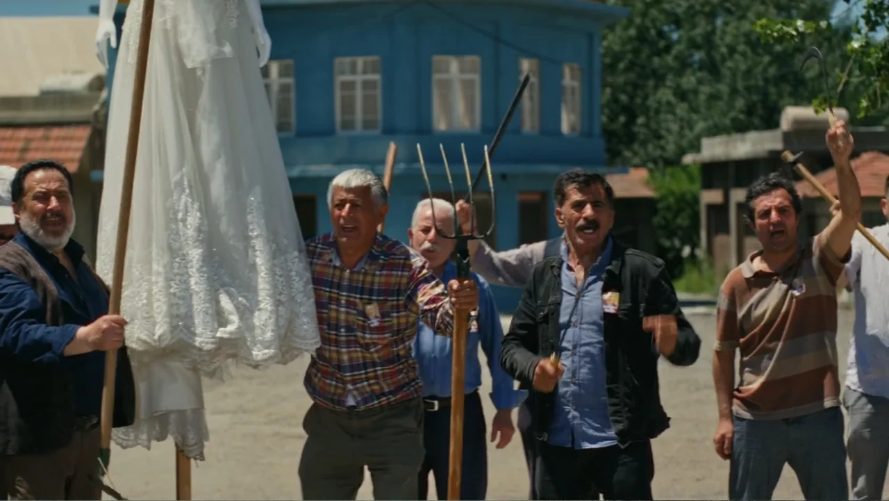 Paranormal Cuma (2024) movie still: medium — A group of men stand in a dusty exterior space with a blue building behind. The men hold various farm…