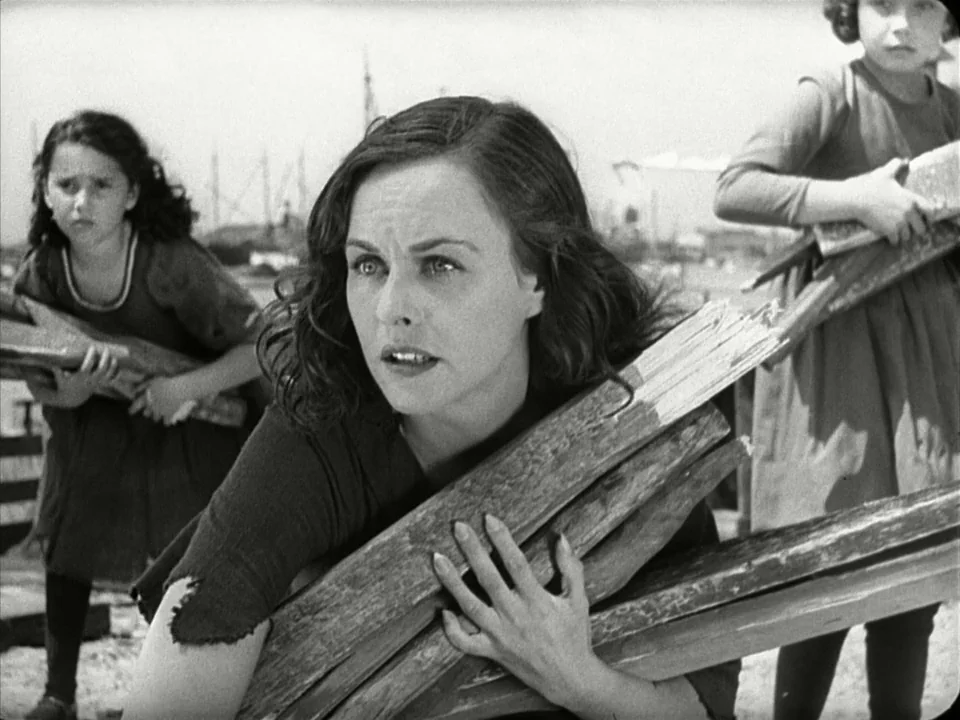 Modern Times (1936) movie still: medium — Three females stand on a harbor dock clutching bundles of rough wooden planks. The central woman stares…
