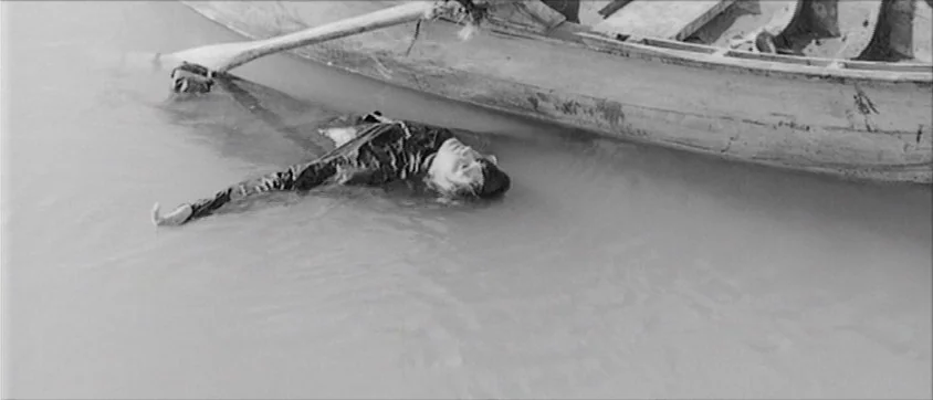 I Vampiri (1957) movie still: wide — A body lies partially submerged in shallow river water next to a wooden rowboat. The person rests supine on the…