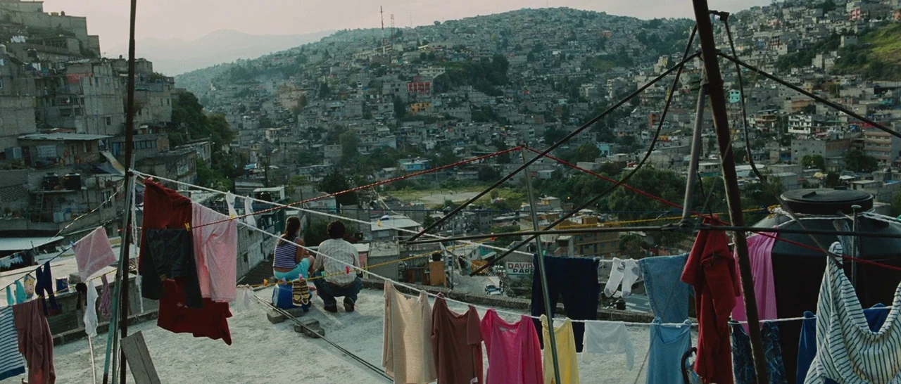 Sin Nombre (2009) movie still: wide — A sprawling neighborhood of concrete houses climbs up a steep hillside in Mexico. A man and woman sit on a…