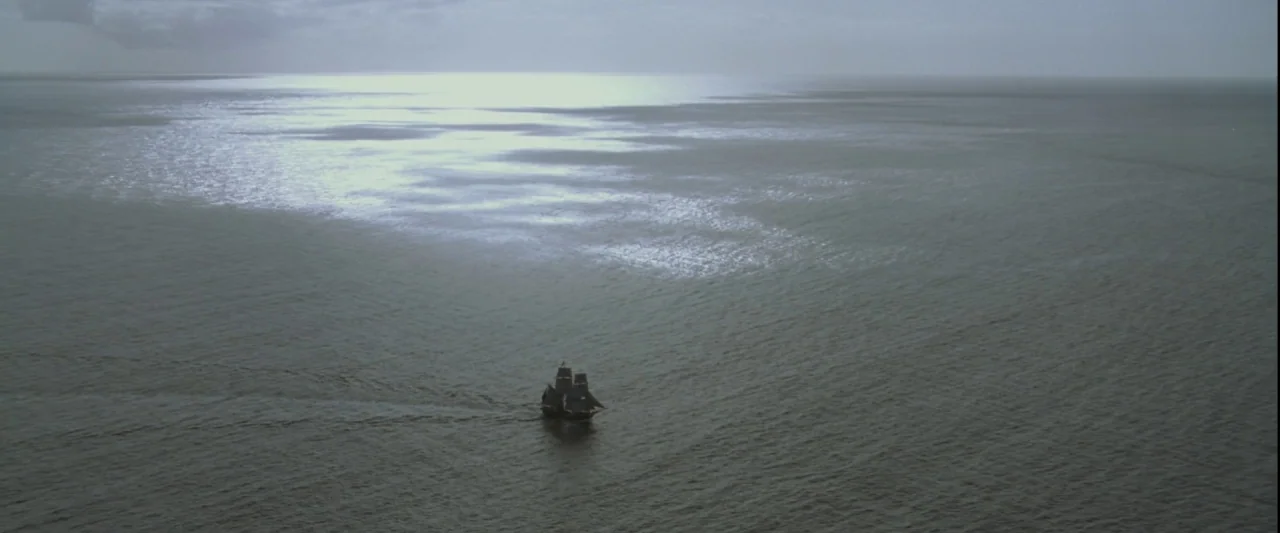 Pirates of the Caribbean: The Curse of the Black Pearl movie still: wide — A vast expanse of calm dark water stretches under a clouded overcast sky.…