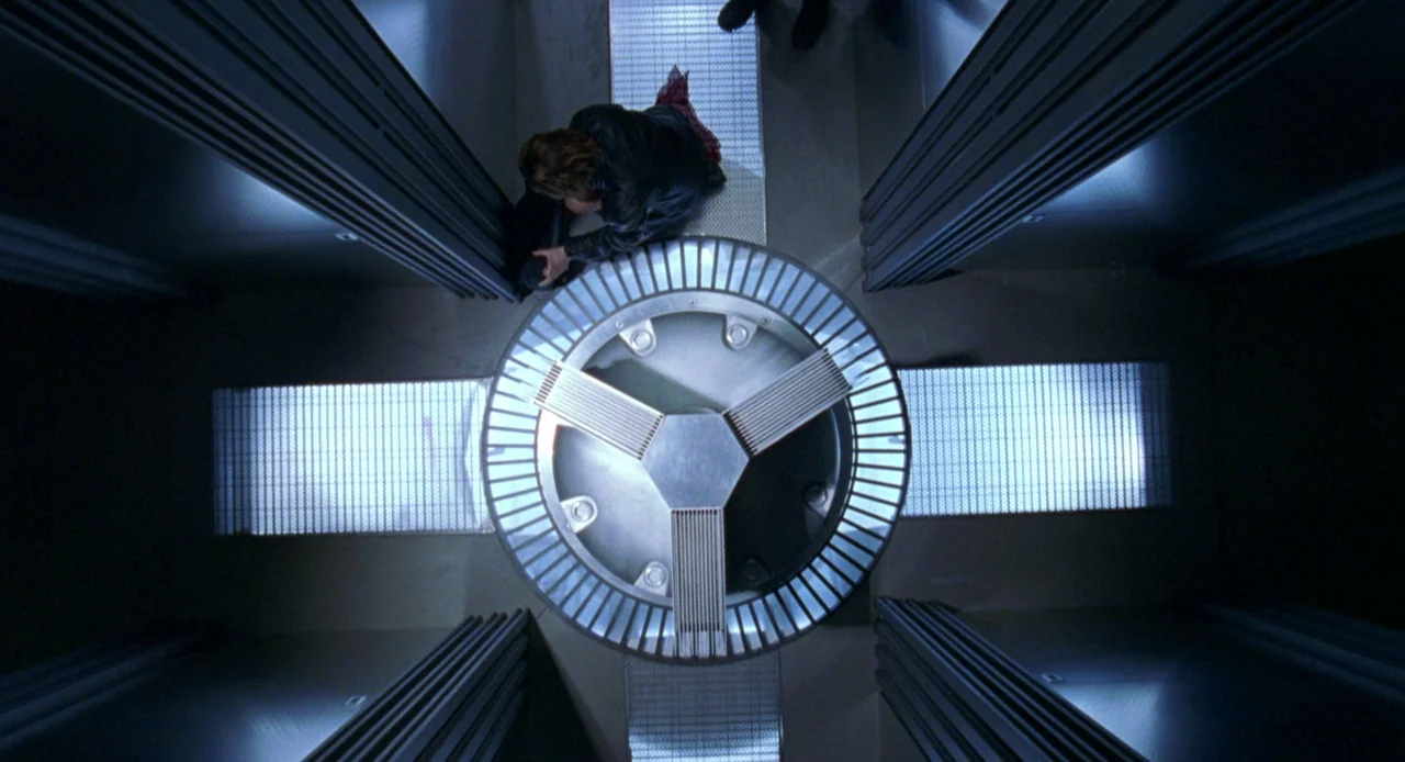 Resident Evil (2002) movie still: wide — An industrial facility features a large circular metal grate assembly set into the floor. A person crouches…