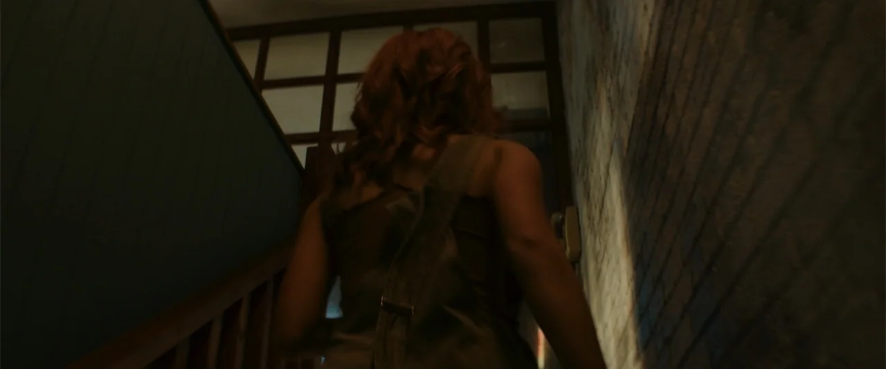 Maintenance Required (2025) movie still: medium — A dimly lit staircase interior with a textured brick wall and wooden railings. A woman in a green…