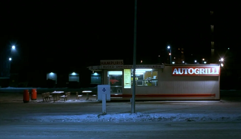 Night on Earth (1991) movie still: wide — A roadside food kiosk sits illuminated at night in a dark, snow-covered parking area. The rectangular…