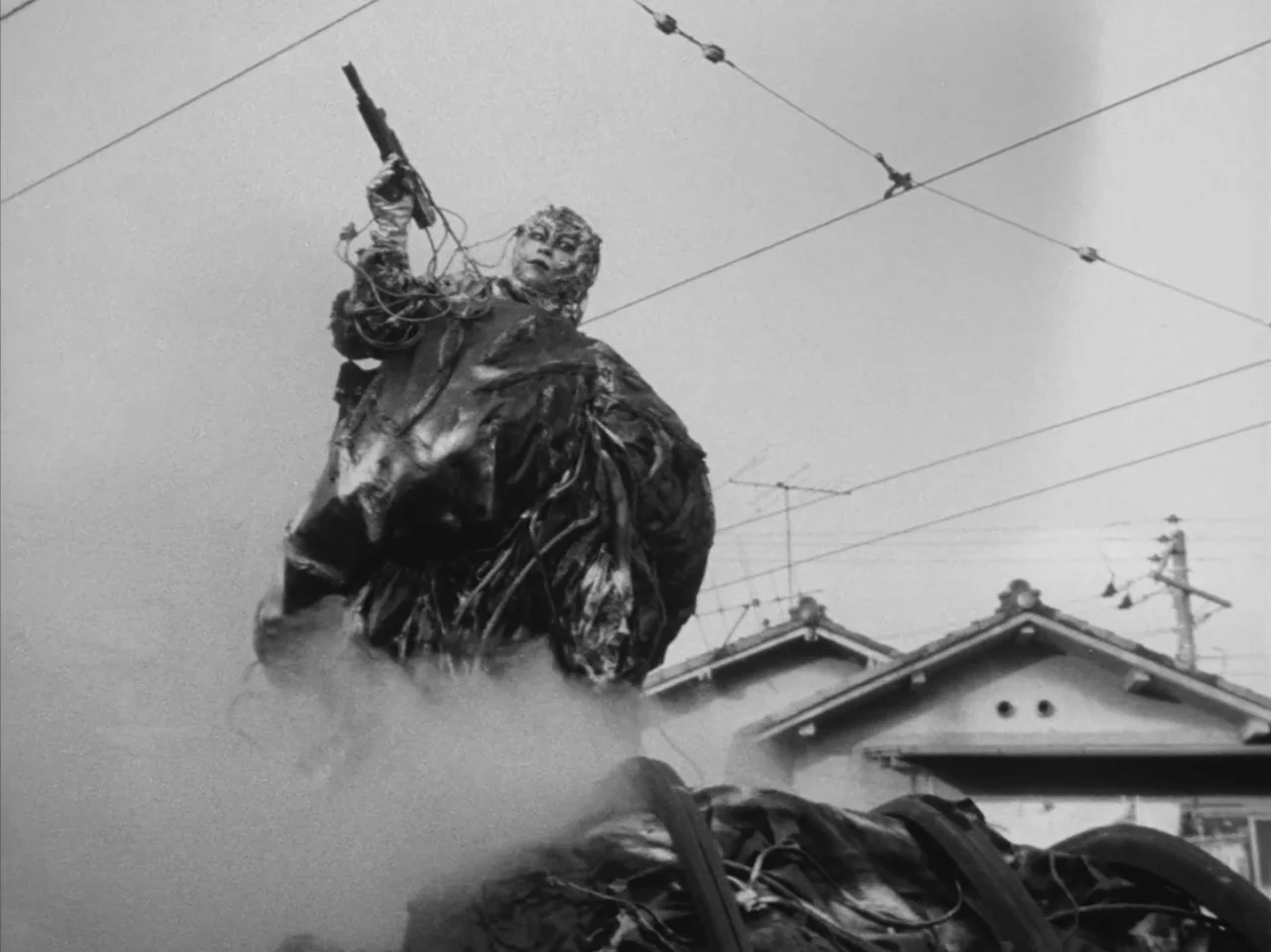 Tetsuo: The Iron Man (1989) movie still: fullbody — A cyborg-like figure stands amid smoke in a Japanese suburban neighborhood. Overhead electrical…