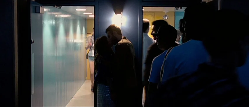 This Movie Is Broken (2010) movie still: medium — A dimly lit indoor hallway with light coming from a doorway at the end. A couple stands in the…