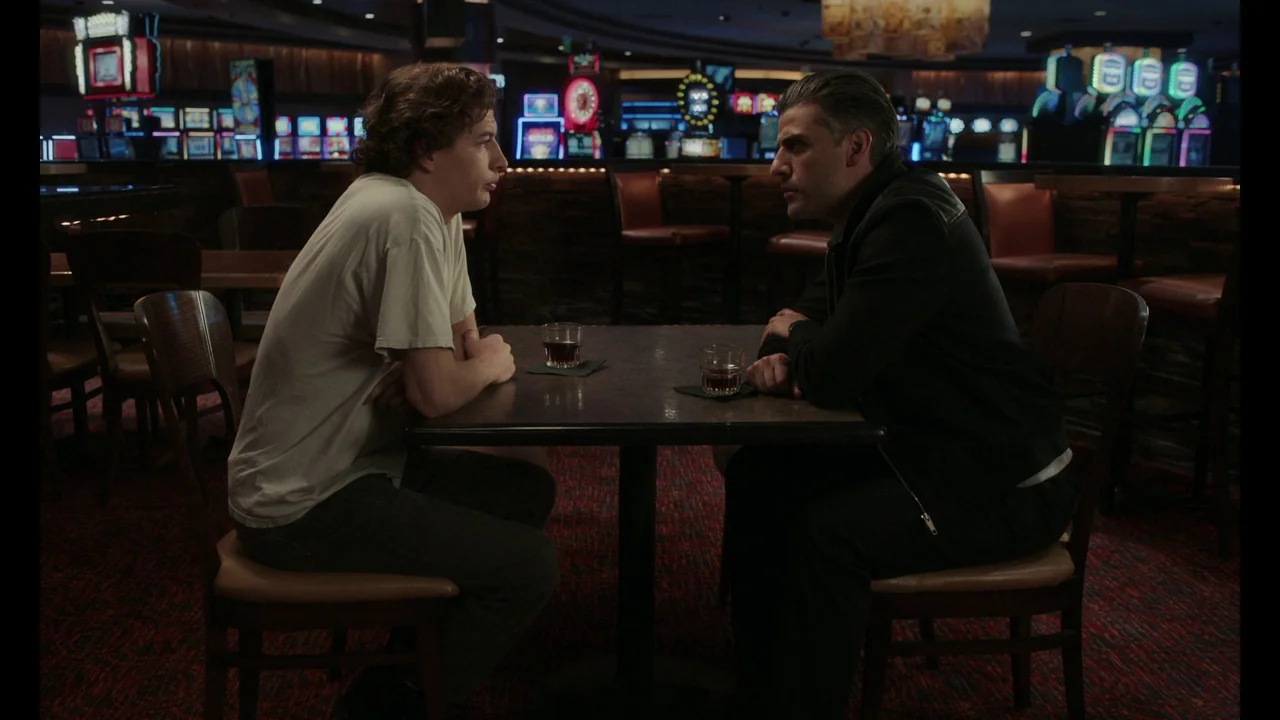 The Card Counter (2021) movie still: fullbody — Two men sit at a square table in the dim expanse of a casino floor. They face each other while…