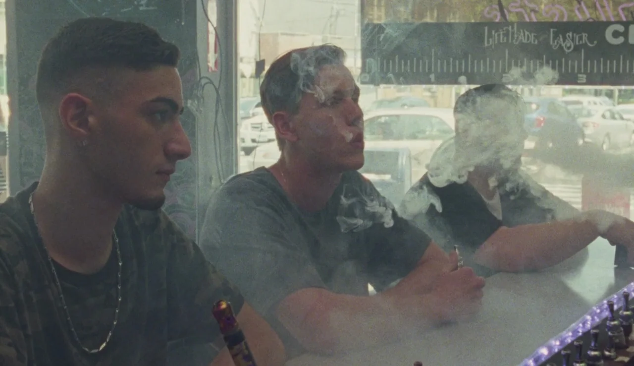 Beach Rats (2017) movie still: medium — Three young men sit behind a long counter in a brightly lit retail space. Thick clouds of vapor fill the air…