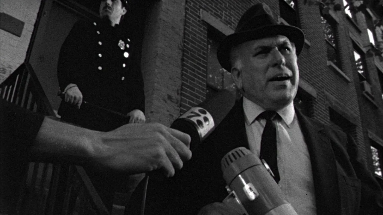 Poison movie still: medium — An older man in a fedora speaks to a reporter outside a brick building. A uniformed police officer stands on the steps…