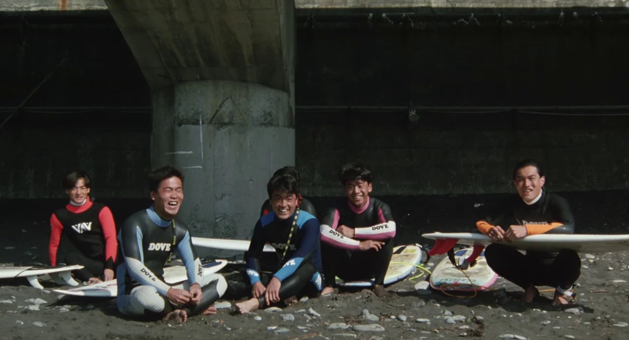 A Scene at the Sea (1991) movie still: fullbody — Five men in wetsuits sit on a pebbled surface beneath a large concrete bridge. The group poses…